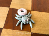 Graue 3D-gedruckte Spinne mit rosa Donut auf dem Rücken